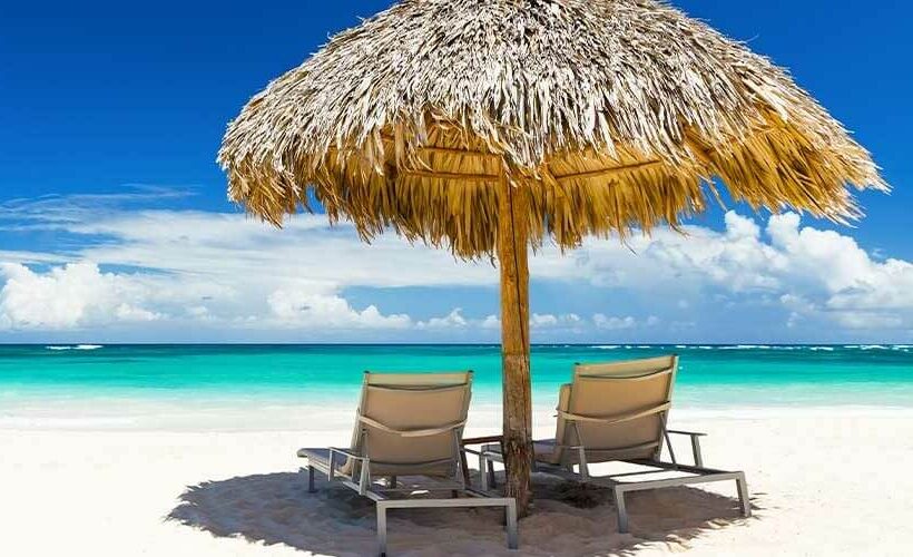 Two lounge chairs sit beneath a thatched umbrella on a sandy beach, inviting relaxation by the ocean. Destinations like this are a key to maximizing your auction's success.